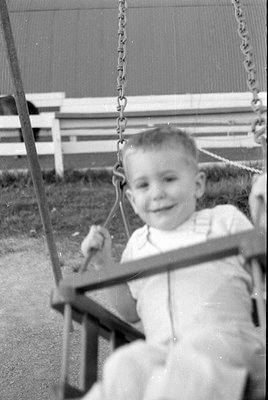 A black and white photograph of a young child sitting on a swing. The child is wearing light-colored overalls and is smiling....