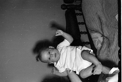 A black and white photograph of an infant lying on their back, wearing a light-colored short-sleeved bodysuit. The child has ...