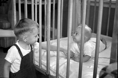 A black and white photograph showing a young child in overalls standing next to a wooden crib. Inside the crib, an infant in ...