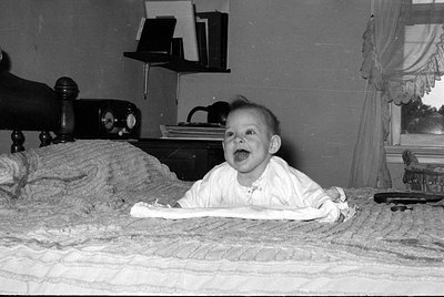 A black and white photograph of a baby lying on a textured bedspread. In the background, a shelf holds books and folders, and...