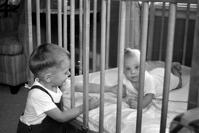 A black and white photograph showing a young child standing outside a wooden crib, looking toward an infant lying inside. The...