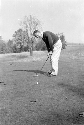 A man in a dark sweater and light-colored trousers stands on a golf course, holding a golf club. A golf ball is positioned on...