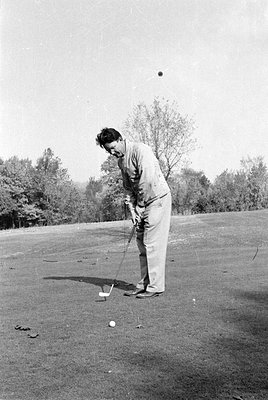 A man in a light-colored jacket and trousers stands on a grassy field, holding a golf club. A golf ball is visible in the air...