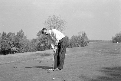 A man in a light-colored long-sleeved shirt and dark trousers stands on a golf course. He is bent over, holding a golf club p...
