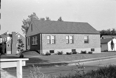 A black and white photograph of a single-story brick house with a gabled roof. The house features several windows and a small...
