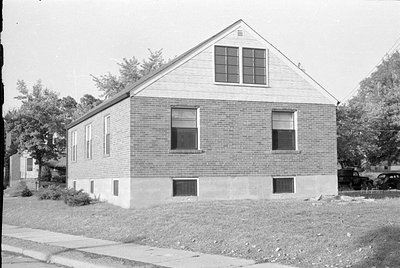 A black and white photograph of a small, single-story brick building with a gabled roof. The upper gable is finished with lig...