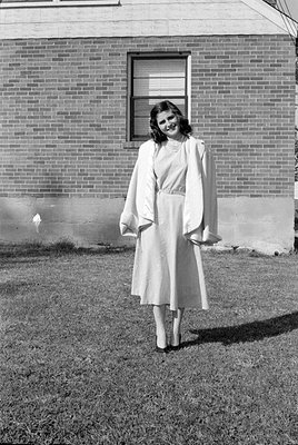 A full-length portrait of a woman standing on a lawn in front of a brick house. She is wearing a light-colored dress, a match...