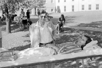 A man in a light-colored suit and sunglasses stands beside a woman lying on a white cot. In the background, other people are ...