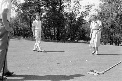 A black and white photograph of three people on a golf course. A man in a white t-shirt and trousers stands in the center, wh...