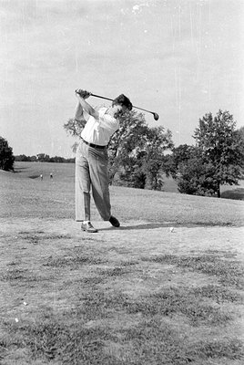 A man in a white short-sleeved shirt and light-colored trousers swings a golf club on a grassy course. A golf ball sits on th...