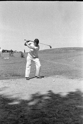 A man in a white short-sleeved shirt and light-colored trousers swings a golf club on a grassy course. A golf ball sits on th...