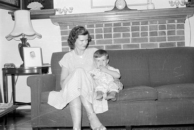 A woman in a striped dress sits on a dark sofa holding a small child. They are positioned in front of a brick fireplace mante...