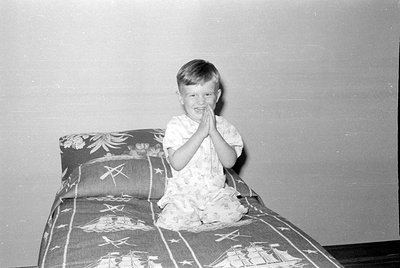 A young child sits cross-legged on a bed, hands pressed together in a prayer-like gesture. The child wears light-colored patt...