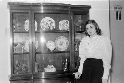A black and white photograph of a woman standing next to a wooden china cabinet. The cabinet contains several decorative plat...
