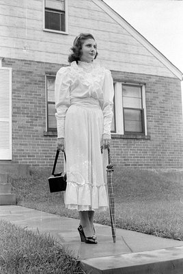 A woman stands on a concrete walkway in front of a brick and siding house. She is wearing a light-colored, long-sleeved dress...