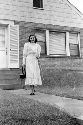 A woman in a light-colored, ruffled ankle-length dress and heels stands on a concrete walkway. She holds a small black handba...