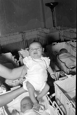 A black and white photograph of a baby held up in a room filled with rows of wicker bassinets. Other infants are visible slee...