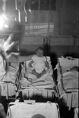 A black and white photograph of an infant in a white garment sitting in a metal hospital bassinet. An arm reaches in from the...
