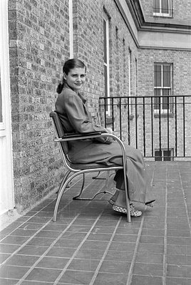A black and white photograph of a woman sitting in a metal-framed chair on a tiled terrace. She is wearing a light-colored su...