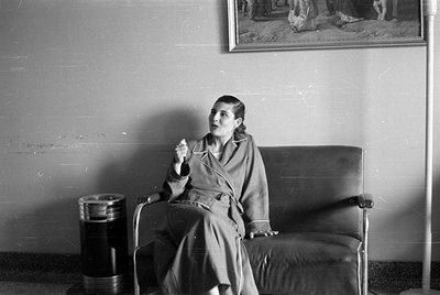 A woman wearing a light-colored robe sits on a dark leather sofa. She is captured mid-speech with one hand raised. A cylindri...