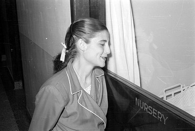 A black and white photograph of a woman in profile, wearing a light-colored jacket with white piping. She is standing next to...