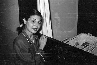 A black and white photograph of a woman in a dark, collared garment looking toward the camera. To her right, a baby lies in a...