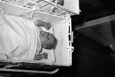 A black and white photograph of an infant lying in a white, quilted bassinet. The baby is wrapped in a light-colored blanket ...