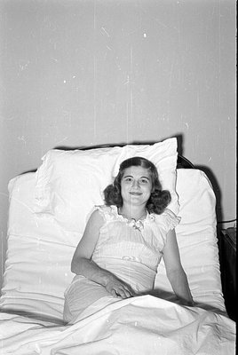 A black and white photograph of a woman with curly hair reclining against large white pillows. She is wearing a light-colored...