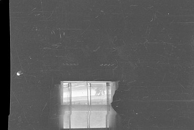 A heavily damaged black and white negative. A small, rectangular section of a window with vertical panes is visible through a...