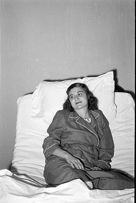 A black and white photograph of a woman reclining against white pillows on a bed. She is wearing dark-colored pajamas with li...