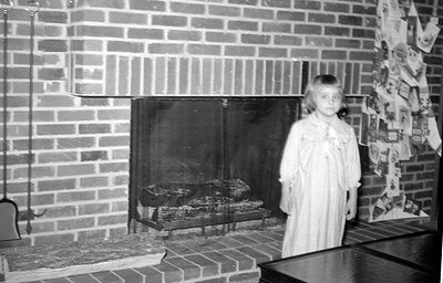 A young girl in a light-colored, long-sleeved dress stands beside a brick fireplace. The fireplace has a black screen and log...