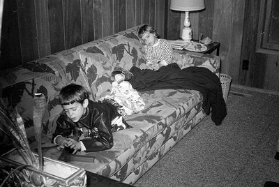 Two children are on a floral-patterned sofa in a room with wood-paneled walls. One child is lying across the back of the sofa...