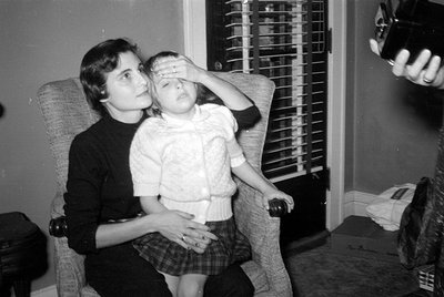 Black and white photograph of a woman and a young child sitting in a textured armchair. The child has one hand over their eye...