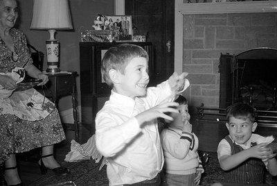 A black and white indoor photograph showing three young boys and a woman. One boy in a white shirt is captured in motion with...