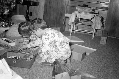 Two children in patterned pajamas sit on a carpeted floor surrounded by open gift boxes and wrapping paper. In the background...