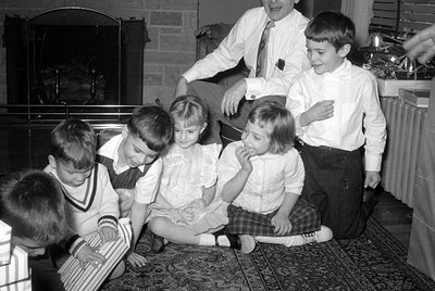 A black and white photograph of four children and one man in a living room. The children are sitting on a patterned rug in fr...