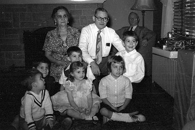 A black and white group portrait of three adults and four children indoors. The adults include a woman in a floral dress and ...