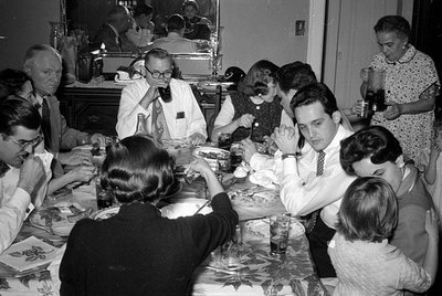 A group of people are seated around a dining table covered with a patterned cloth. Men are wearing white shirts and ties, and...