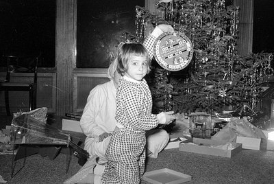 A young child in patterned pajamas stands before a decorated Christmas tree, holding up a circular 'Spelling and Counting Boa...