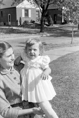 A black and white photograph of a woman and a young child outdoors. The child wears a light-colored, ruffled fancy dress and ...