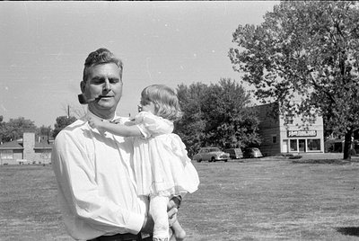 A black and white photograph of a man with a pipe in his mouth holding a young child in a white dress. They are outdoors on a...