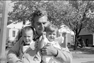 A black and white photograph of a man holding a young girl and a young boy. The boy is wearing a light-colored suit jacket an...