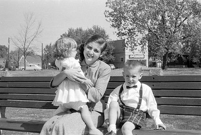 A woman and two children sit on a wooden park bench. The woman holds a young child in a white dress, while a boy in a white s...