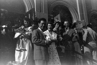 A black and white photograph of a group of people standing in front of a stone arched entrance. A woman in a white dress hold...