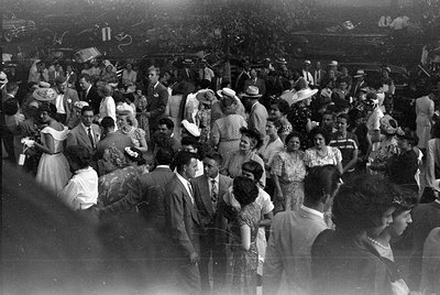 A large crowd of people gathered outdoors, many wearing formal attire including suits, floral dresses, and wide-brimmed hats....