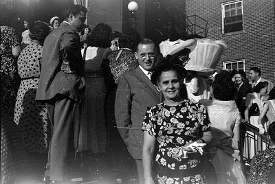 A group of people gathered outdoors against a brick building. A man in a suit and a woman in a floral print dress stand in th...