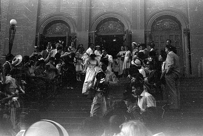 A black and white photograph of a crowd gathered on the steps of a building with three large arched doorways. A person in a w...