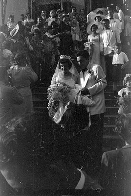 A black and white photograph of a couple in wedding attire descending a set of stairs. The woman wears a white gown, veil, an...