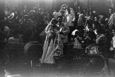A black and white photograph of a couple in wedding attire standing in a crowded area. The woman wears a white gown and veil,...