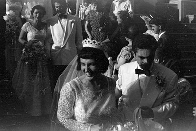 Black and white photograph of a couple in wedding attire in the foreground, with other formally dressed guests behind them. T...